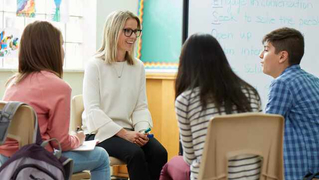 A woman sits in a circle with three students in a classroom, smiling as they engage in a group discussion.