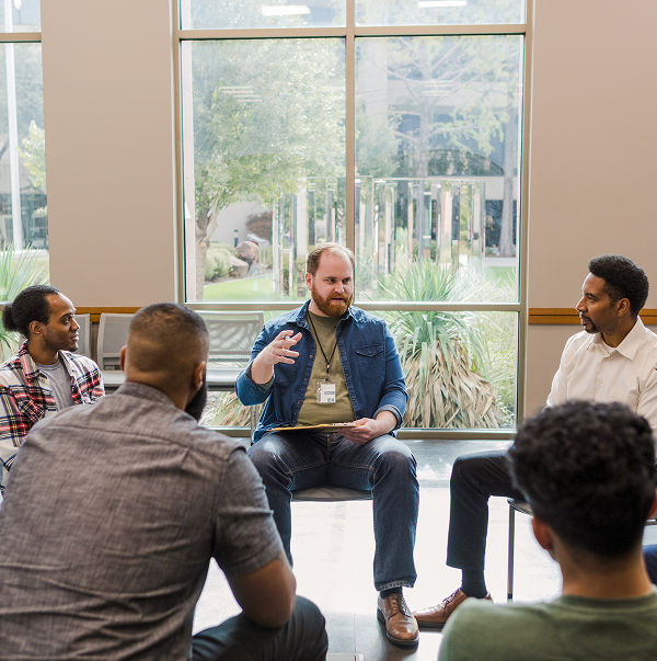A group of men sit in a circle during a discussion, with one man in the center speaking and gesturing with his hands.