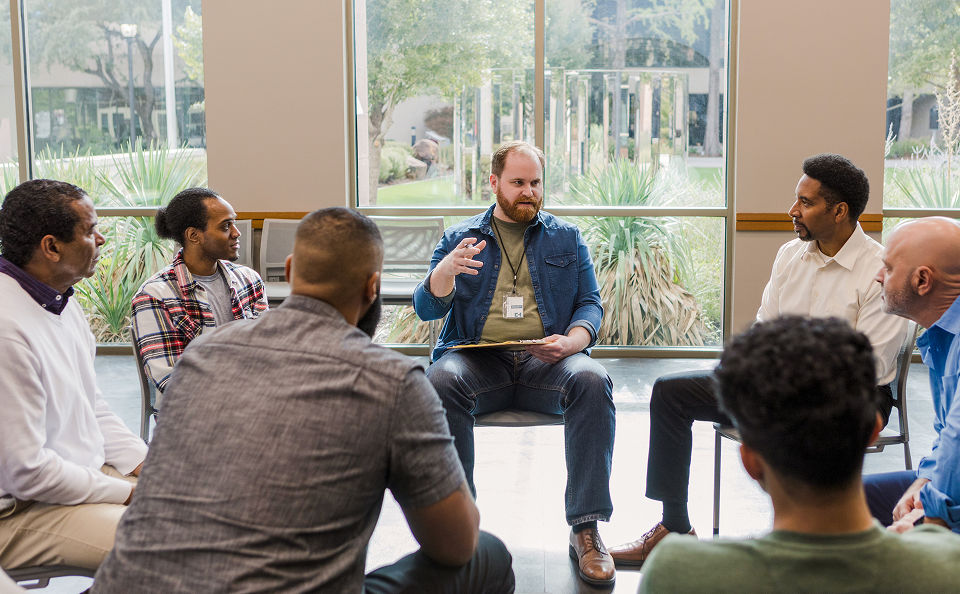 A group of men sit in a circle during a discussion, with one man in the center speaking and gesturing with his hands.