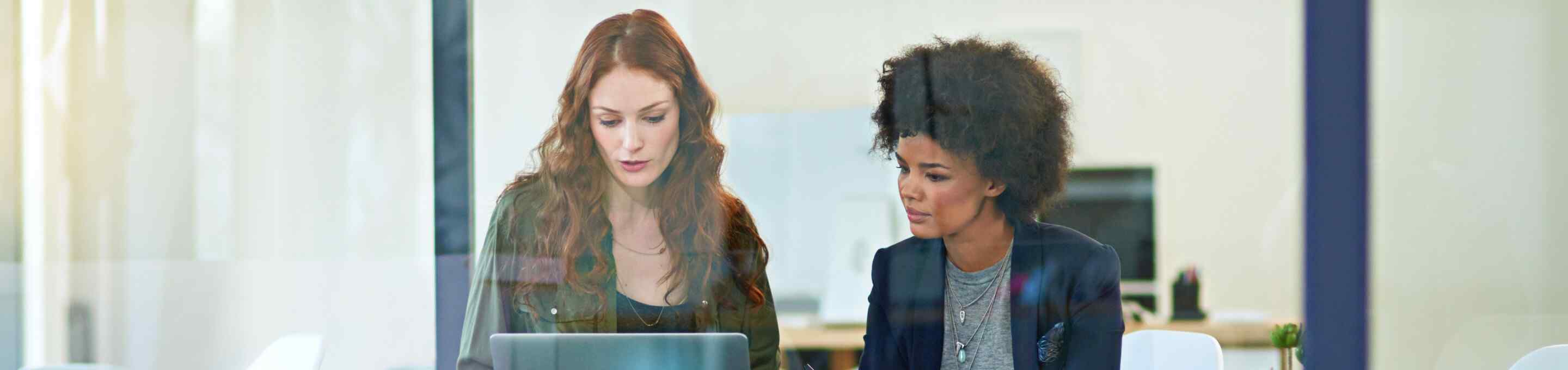 two women talking while looking at a computer from behind a window