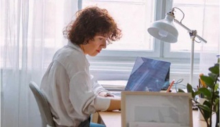 A woman sits at a wooden desk using a laptop in a softly lit room, with a white lamp and houseplants nearby.