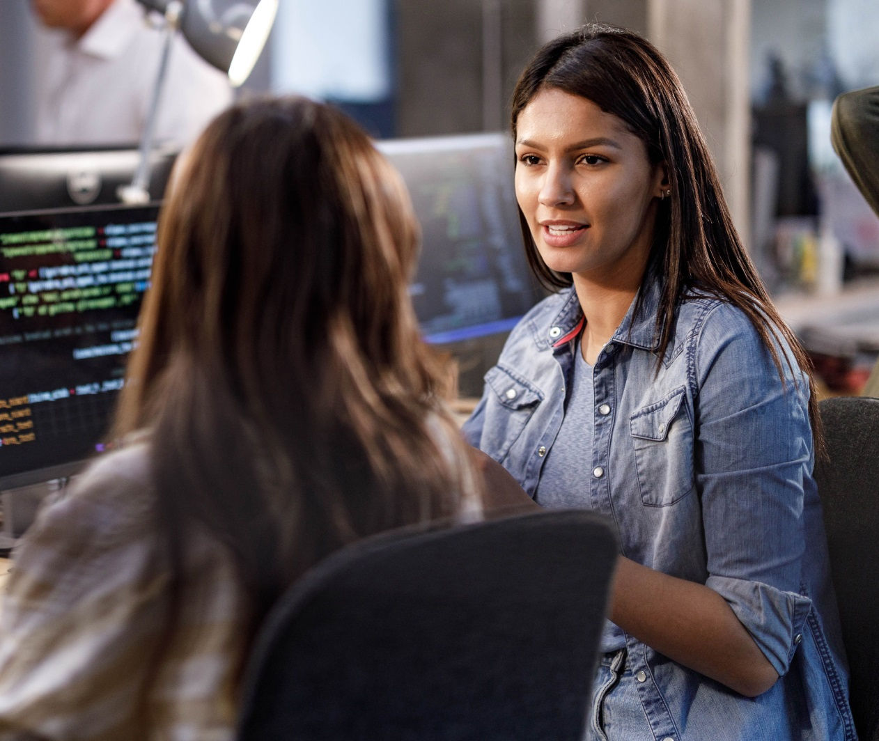 Two women in a modern office discussing code displayed on large monitors