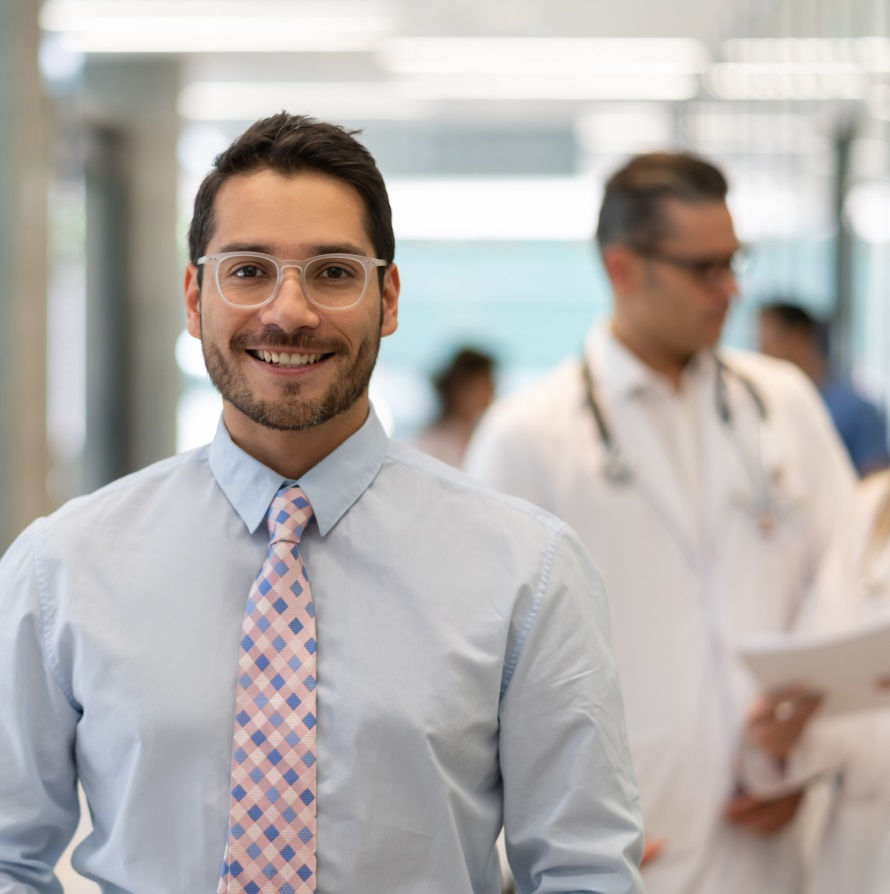 Smiling man wearing glasses, a light blue shirt, and a checkered tie standing in a hospital hallway