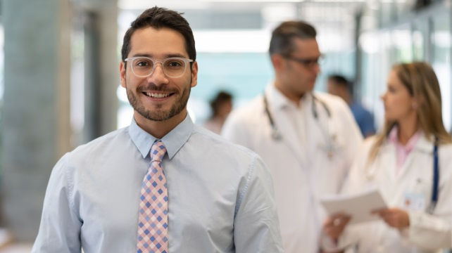 Smiling man wearing glasses, a light blue shirt, and a checkered tie standing in a hospital hallway