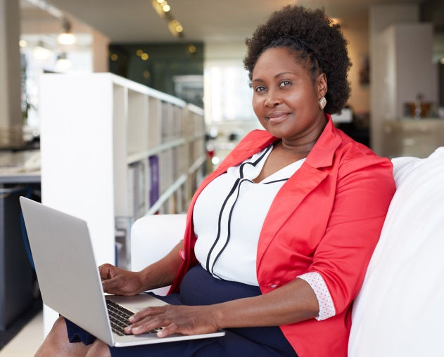 A woman sits on a white couch using a laptop, dressed in a red blazer and white blouse, with bookshelves and modern office decor in the background.