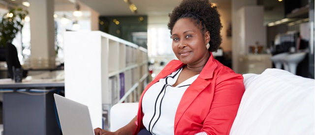 A woman sits on a white couch using a laptop, dressed in a red blazer and white blouse, with bookshelves and modern office decor in the background.