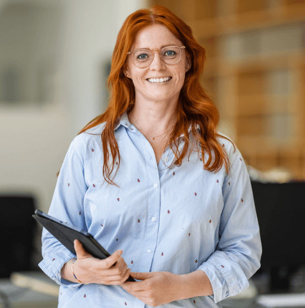 A smiling woman with long red hair and glasses stands indoors holding a tablet, wearing a light blue button-down shirt with small red patterns.