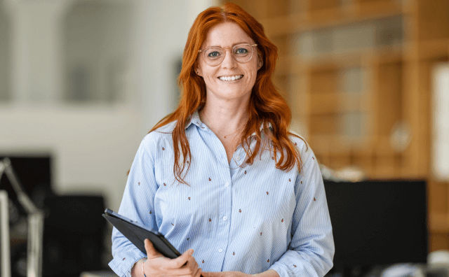 A smiling woman with long red hair and glasses stands indoors holding a tablet, wearing a light blue button-down shirt with small red patterns.